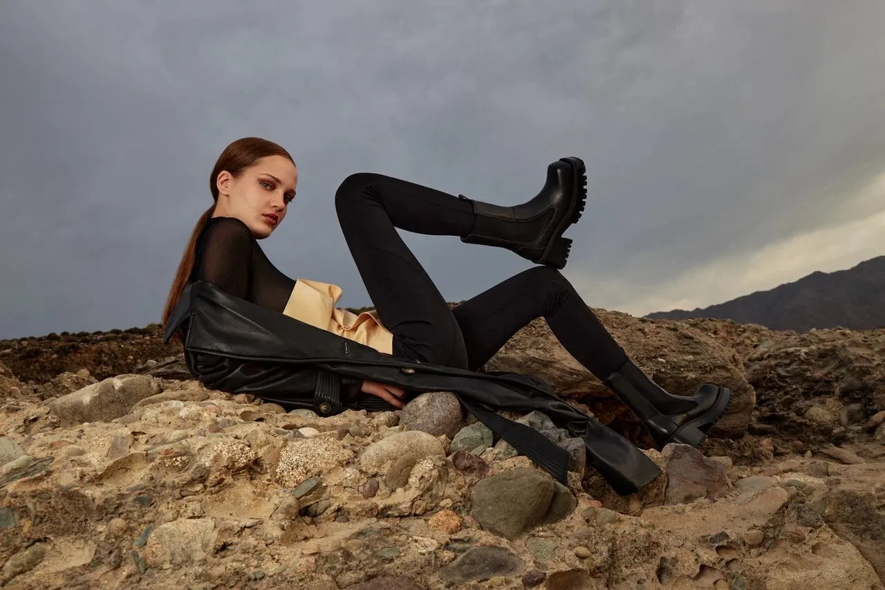 Commercial footwear campaign featuring a model in a black bodysuit and over-the-knee boots, photographed outdoors on a rocky landscape.
